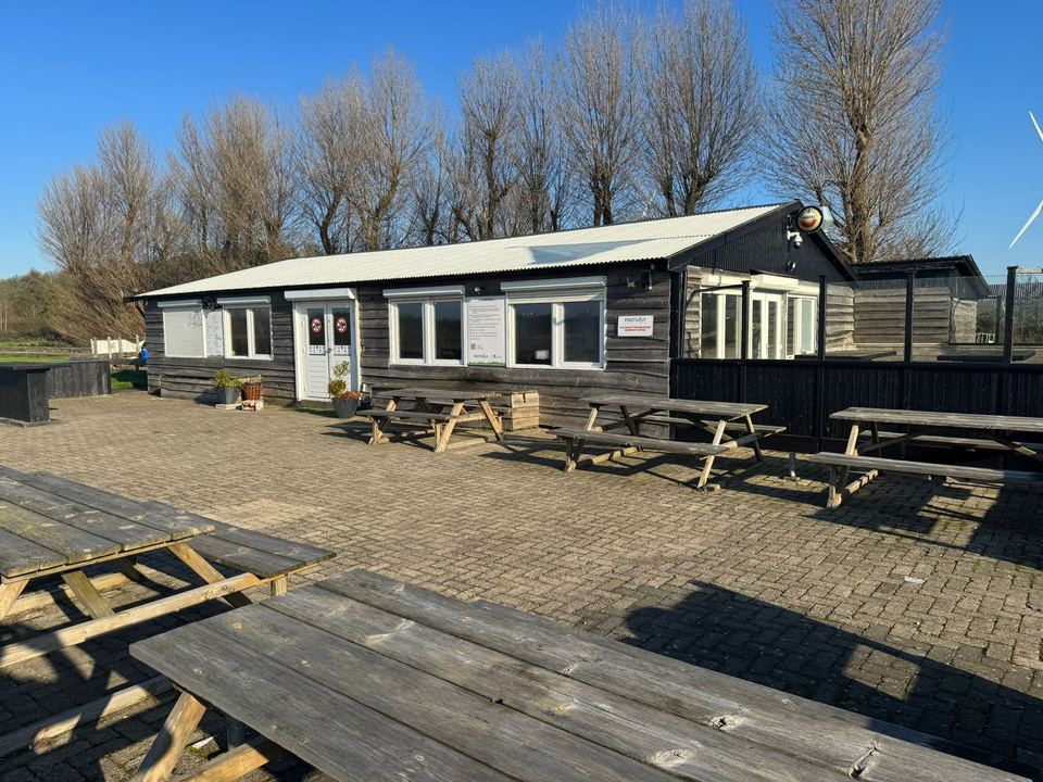Het zwarte houten clubhuis van FG Zaalverhuur met terras en picknicktafels in de zon